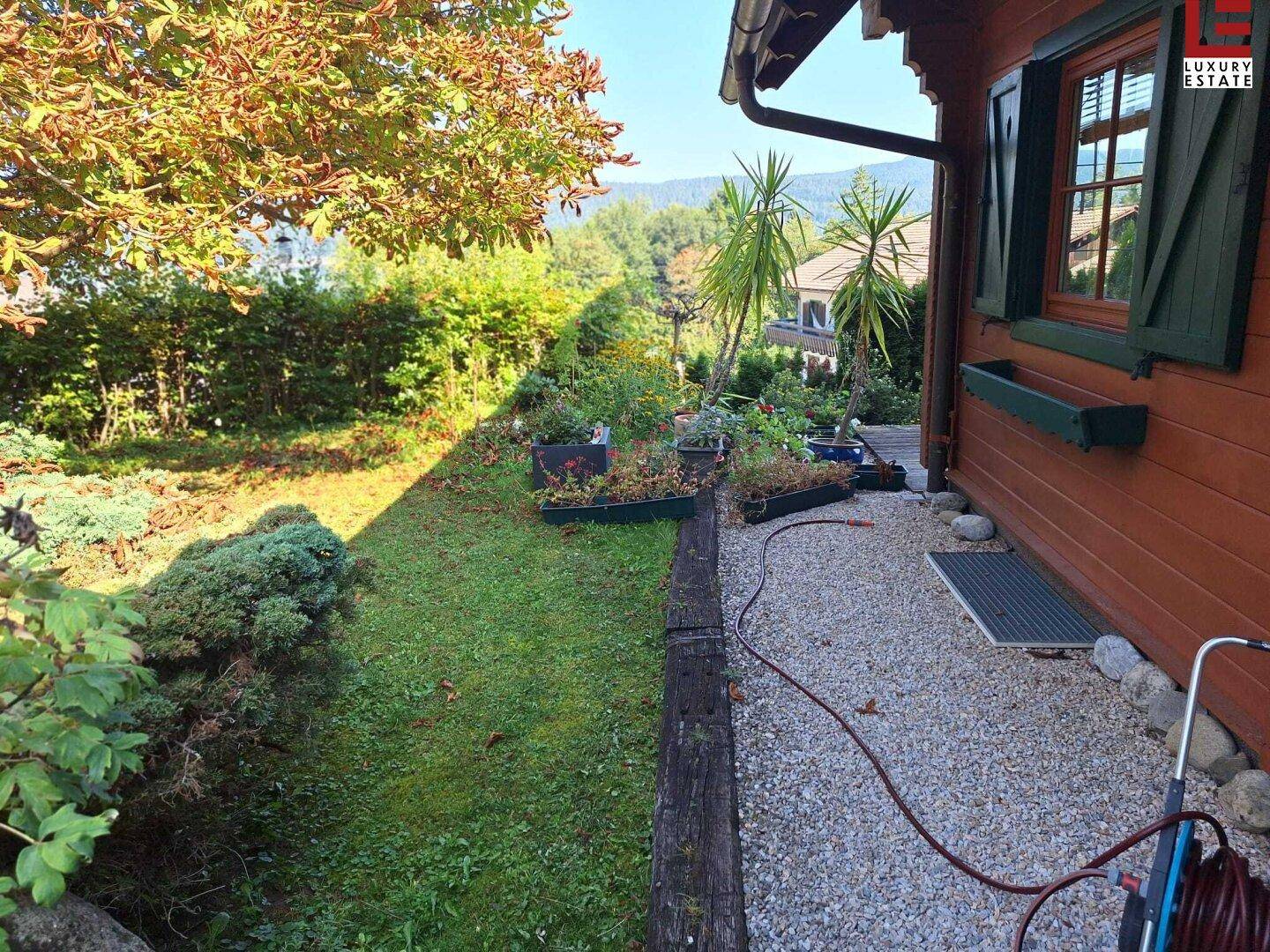 Seitlicher Gartenbereich mit Kiesweg, Pflanzenbeeten und Blick auf die umliegende Natur.