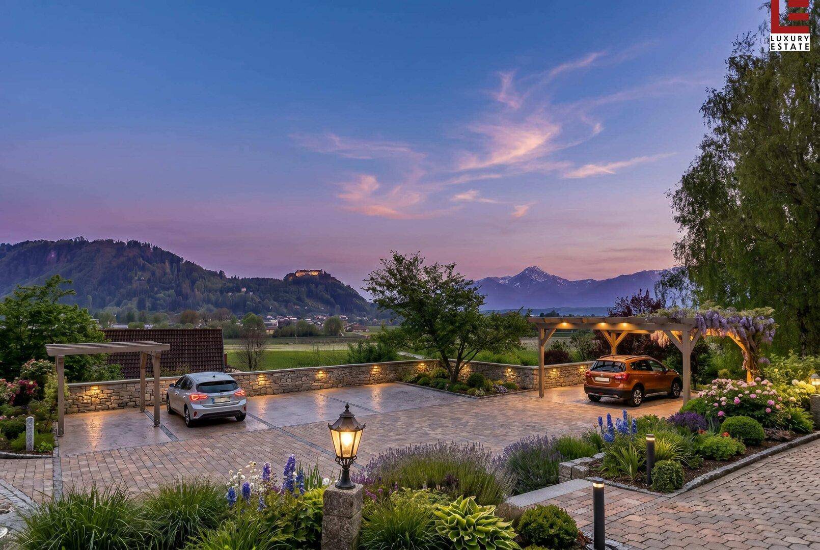 Beleuchteter Parkplatz mit Carports, gepflegten Blumenbeeten und malerischem Bergblick bei Sonnenuntergang.