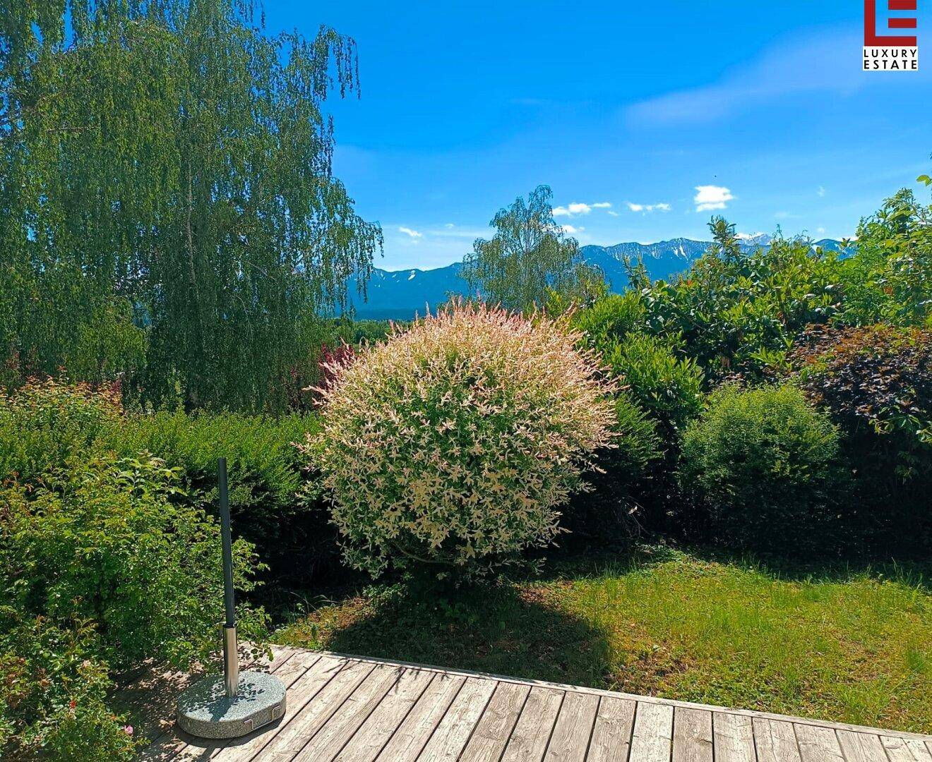 Holzterrasse mit Blick auf einen gepflegten Garten und üppige Vegetation unter blauem Himmel.