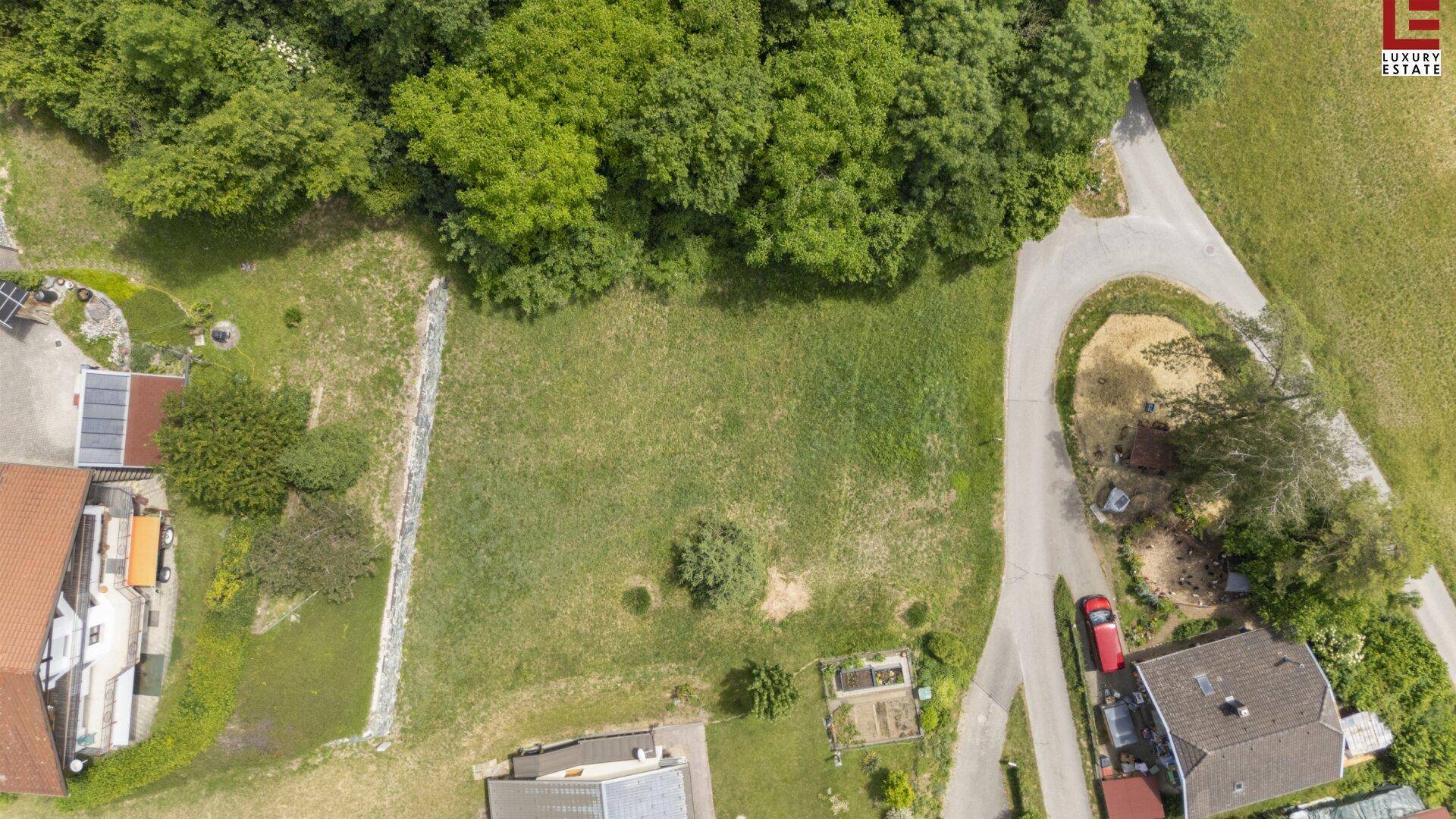 Blick von oben auf ein grünes, unbebautes Grundstück, umgeben von Bäumen und angrenzenden Wohnhäusern.