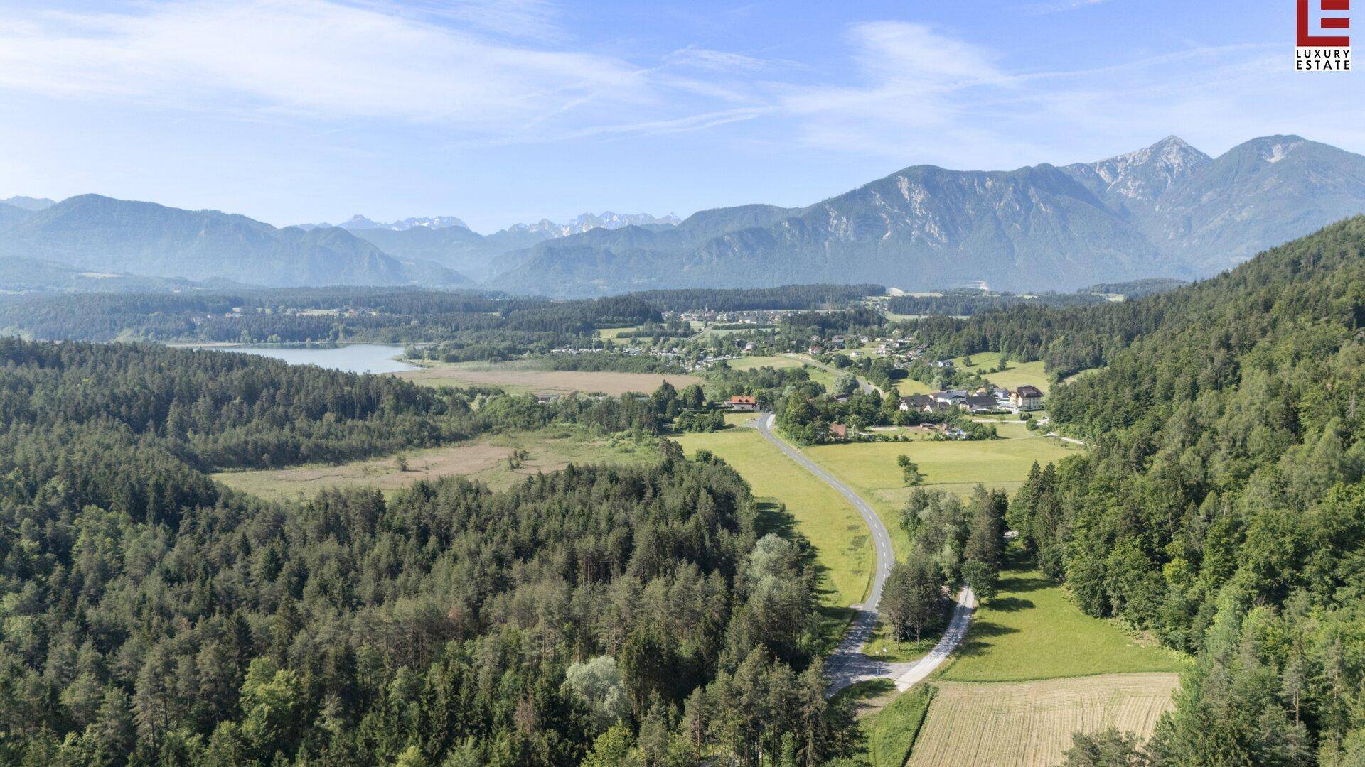 Weitläufige Landschaftsaufnahme mit Wäldern, Feldern, einem See und majestätischen Bergen im Hintergrund.