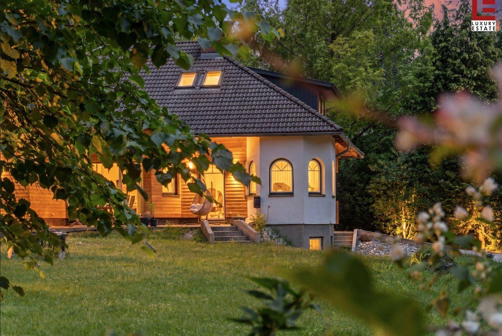 Charmantes Haus in der Abenddämmerung, umgeben von einem gepflegten Rasen und beleuchteten Bäumen.