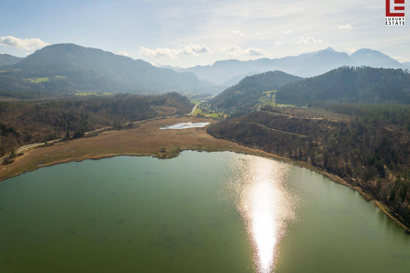 Luftaufnahme eines Sees mit Sonnenreflexion, umgeben von bewaldeten Hügeln und fernen Bergen.
