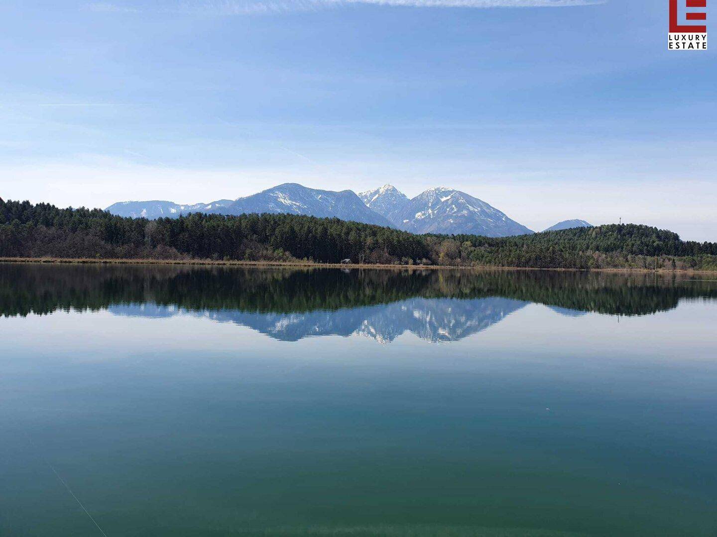 Ruhiger See, der die schneebedeckten Berge und grünen Wälder unter blauem Himmel widerspiegelt.