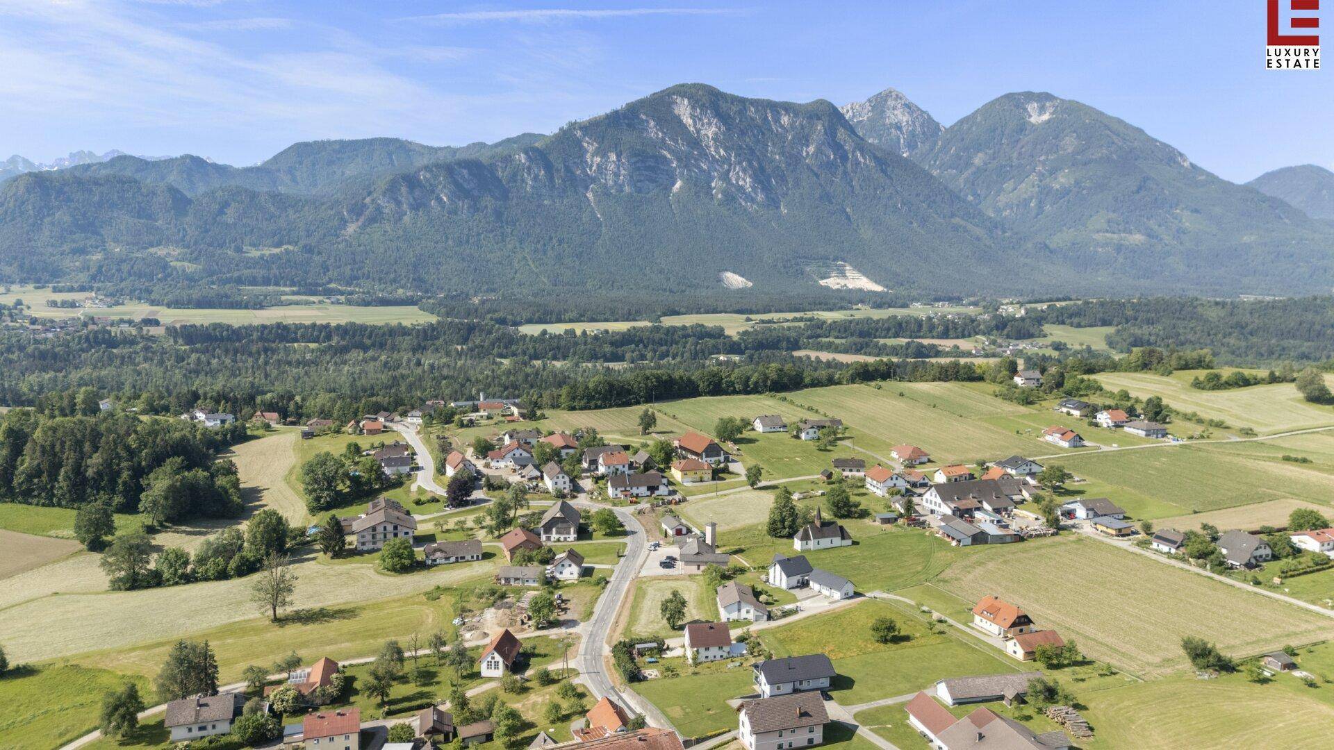 Luftaufnahme eines ländlichen Dorfes in einem Tal, umgeben von grünen Feldern und hohen Bergen.