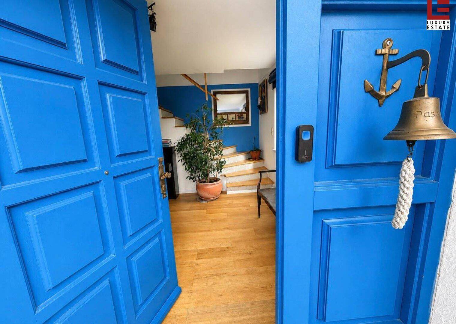 Offene blaue Eingangstür führt in einen hellen Flur mit Holztreppe und Pflanzen.