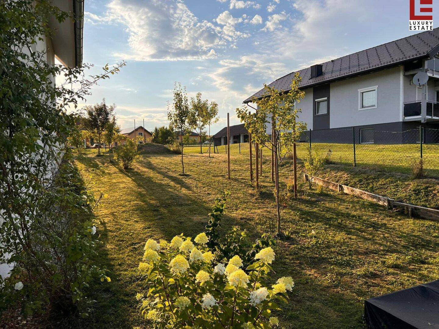 Sonnige Gartenanlage mit blühenden Sträuchern, jungen Bäumen und gepflegtem Rasen.