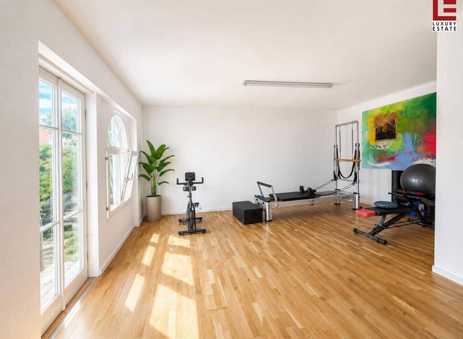 Heller Fitnessraum mit großen Fenstern, Holzboden und verschiedenen Trainingsgeräten.