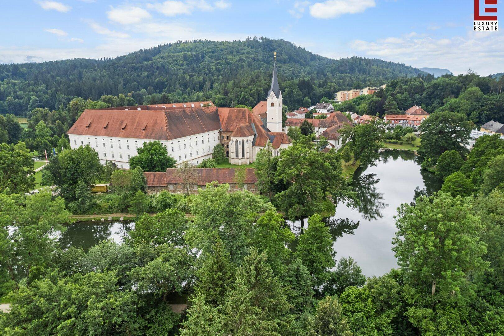 Historisches Klostergebäude mit Kirche, umgeben von üppiger Natur und einem Teich.