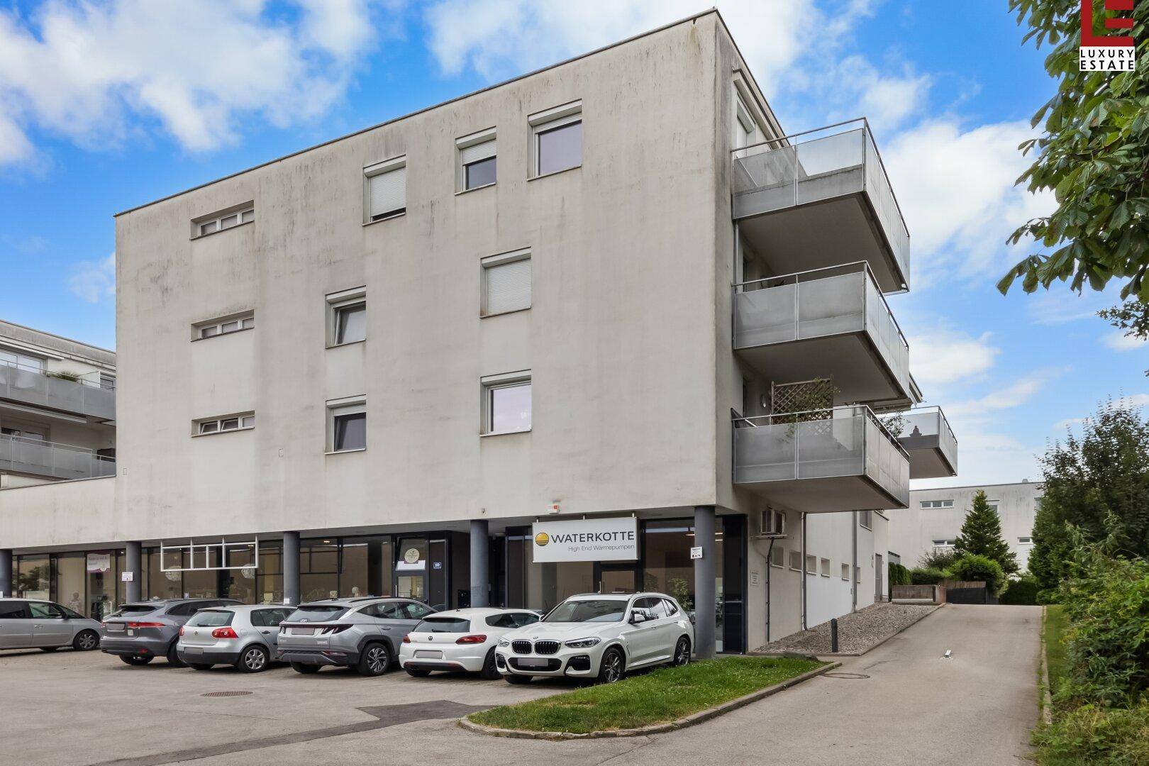 Modernes Mehrfamilienhaus mit Balkonen, großen Fenstern und Parkplätzen vor dem Gebäude.