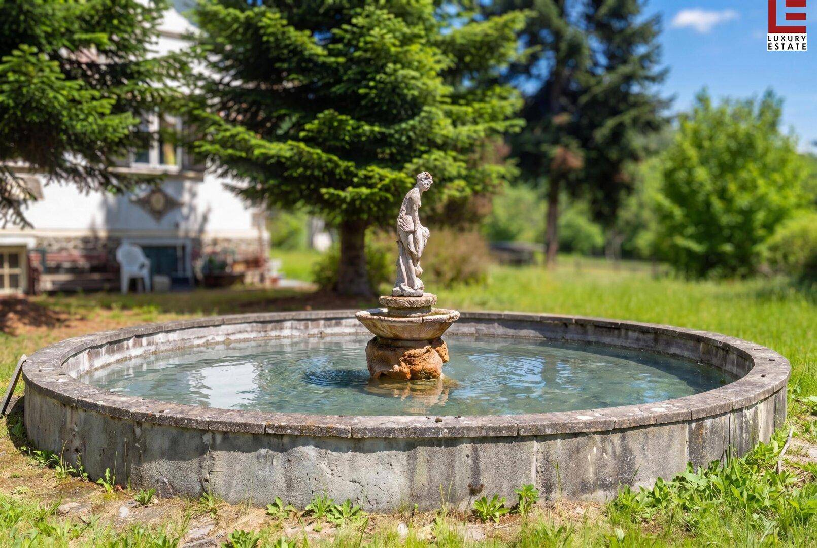 Dekorativer Brunnen im Gartenbereich, umgeben von grüner Wiese und Bäumen.
