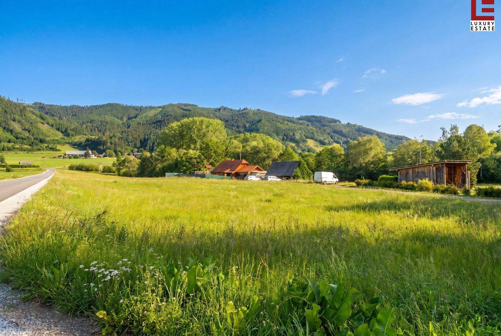 Weitläufige grüne Landschaft mit Hügeln, Bäumen und einigen Häusern in der Ferne.