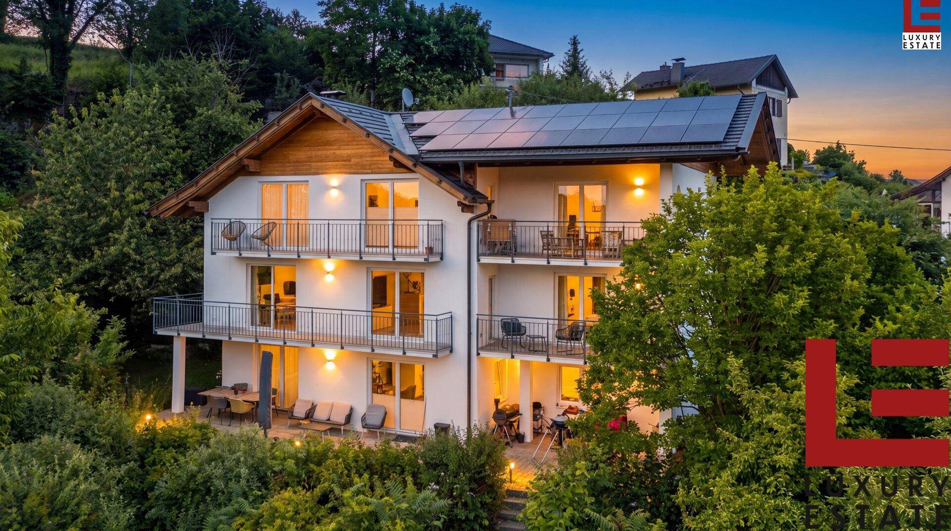 Beleuchtetes Wohnhaus bei Sonnenuntergang mit mehreren Balkonen und Photovoltaikanlage, umgeben von Natur.