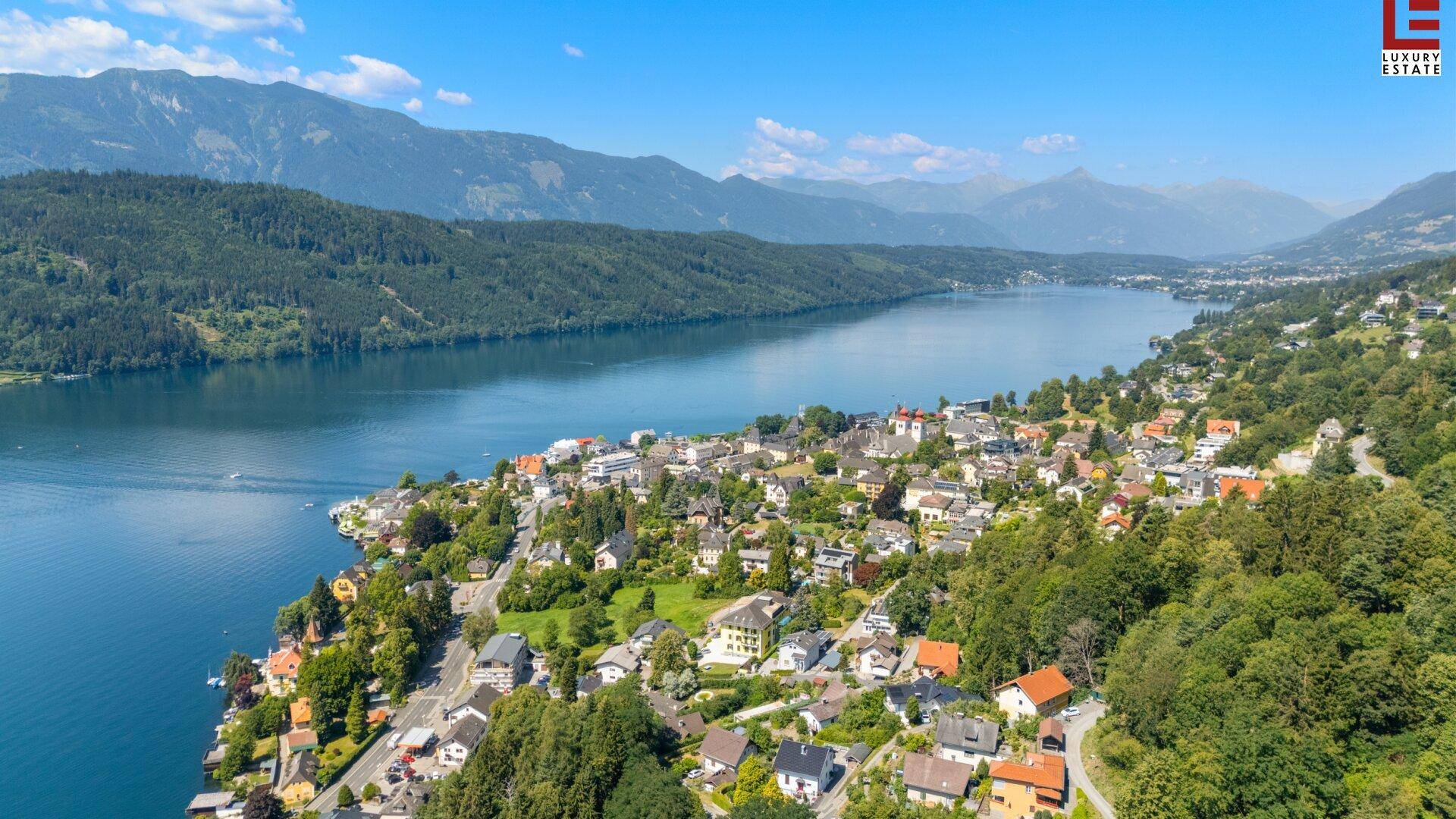 Weitläufige Luftaufnahme einer Stadt am Seeufer, umgeben von Bergen und üppiger Vegetation.