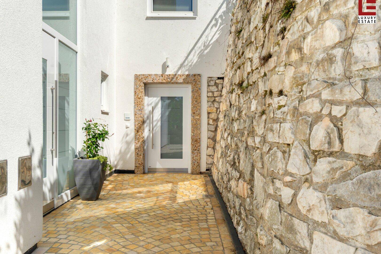 Eingangsbereich des Hauses mit Natursteinmauer und gepflastertem Weg, umgeben von Grün.