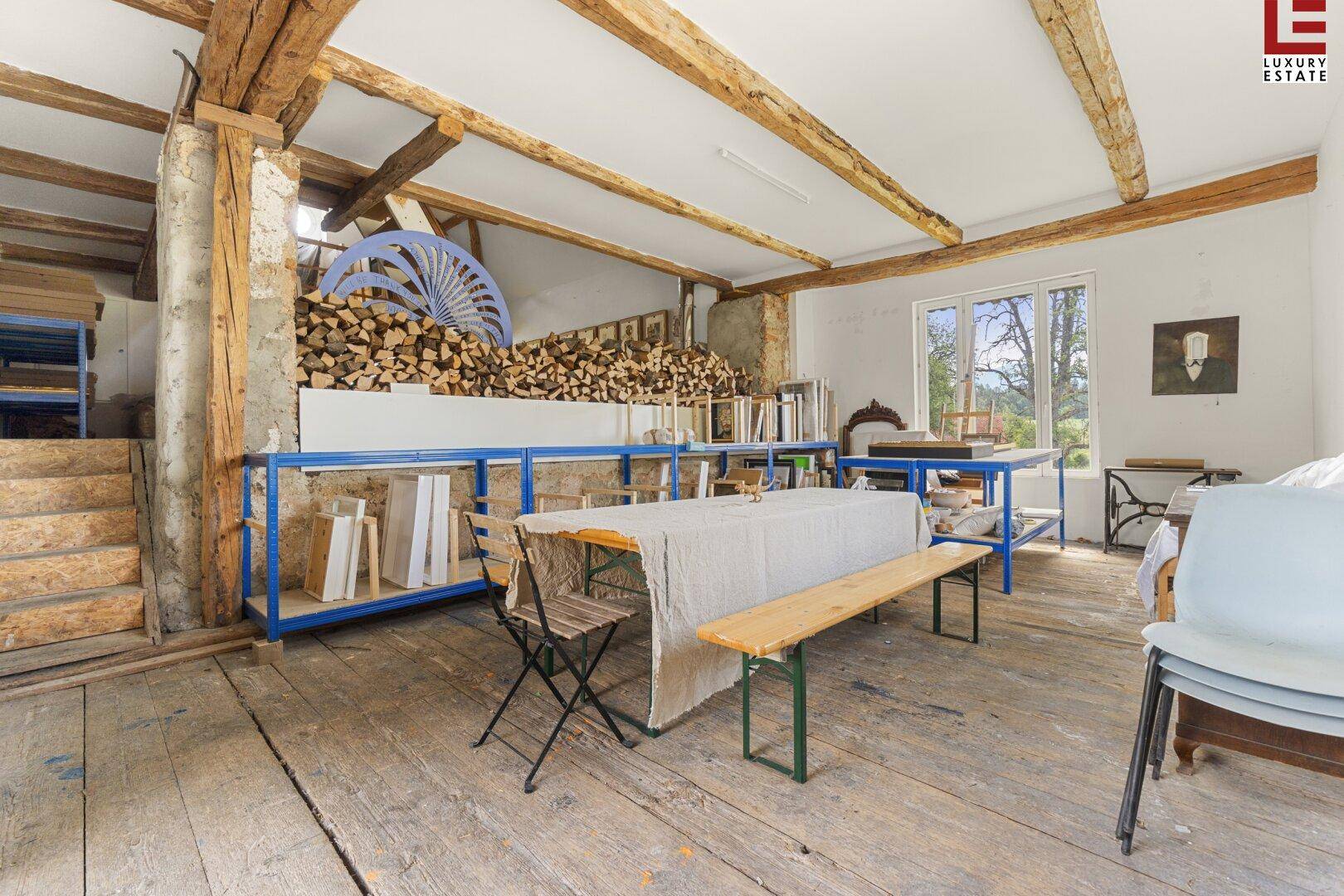 Großer offener Raum mit Holzbalken, Holzdielenboden und einem Stapel Brennholz an der Wand.