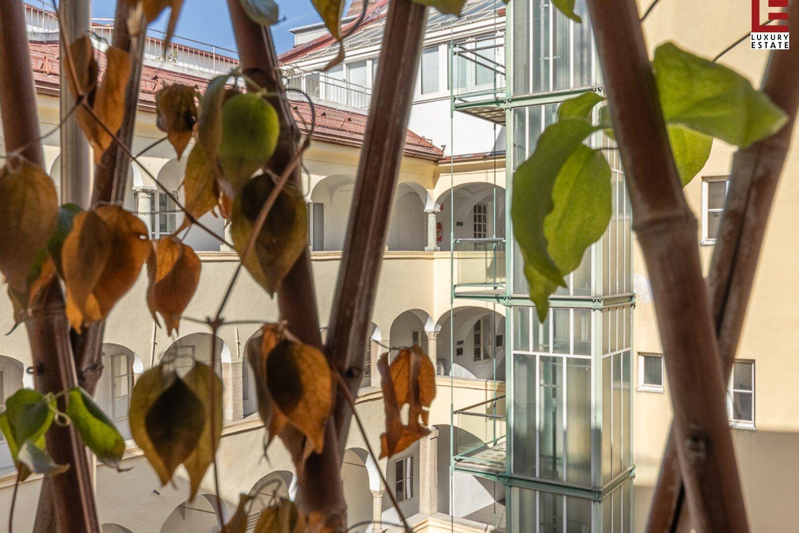 Blick in einen hellen Innenhof mit moderner Glasliftanlage und historischen Arkadengängen.