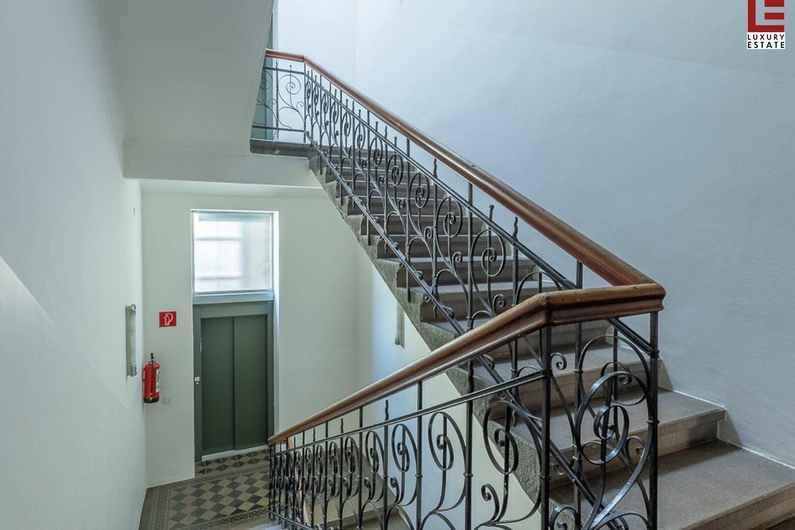 Historisches Treppenhaus mit kunstvollem Geländer und Holzhandlauf, daneben ein Aufzug.