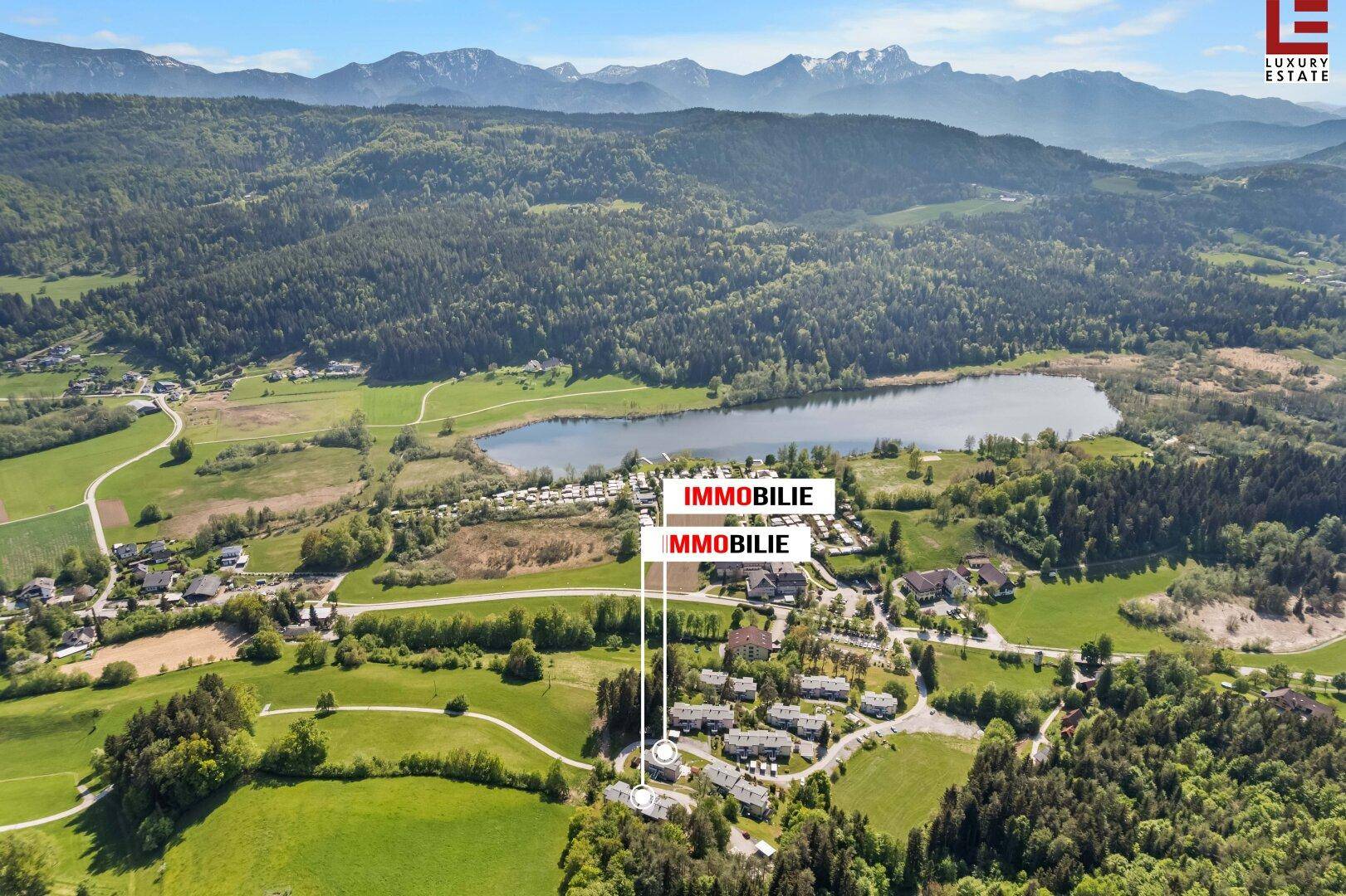 Weitläufige Luftaufnahme einer Wohnsiedlung am Seeufer, umgeben von Wäldern und Bergen, mit markierten Immobilien.