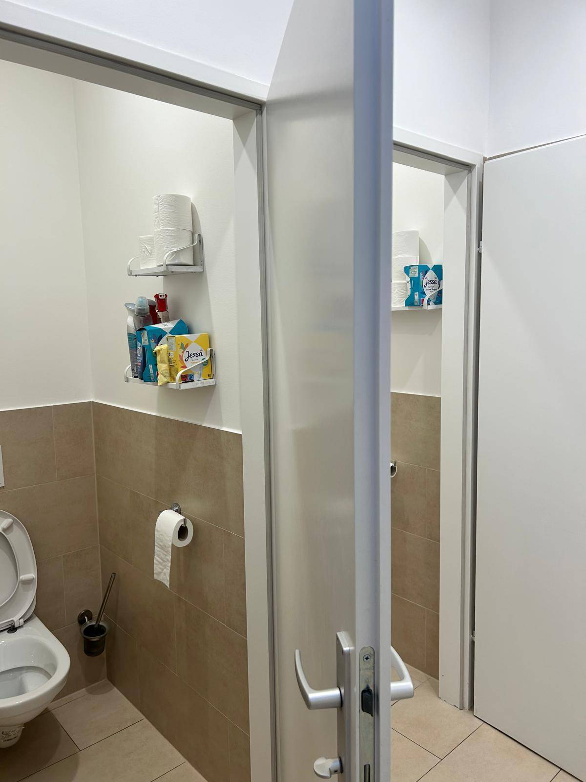 Toilettenbereich mit beigen Fliesen, einer Toilette und Regalen für Hygieneartikel.