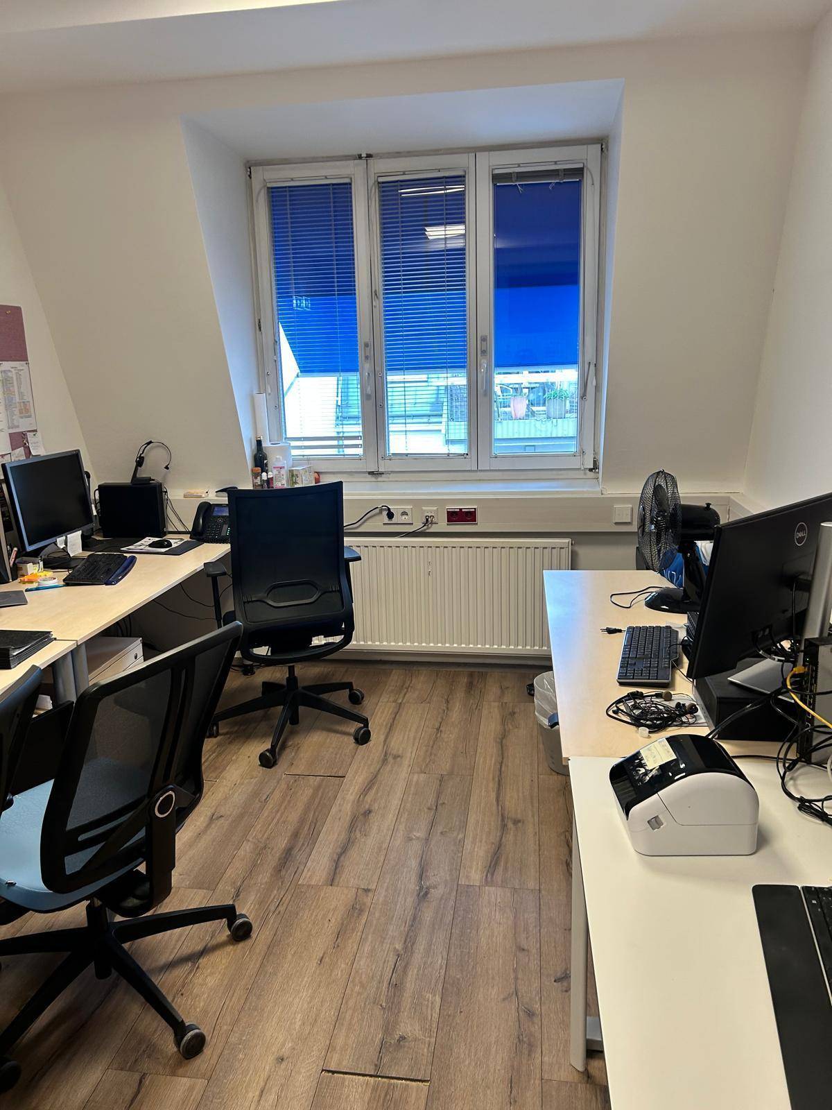 Büro mit mehreren Arbeitsplätzen, blauen Jalousien und einem Ventilator für Belüftung.