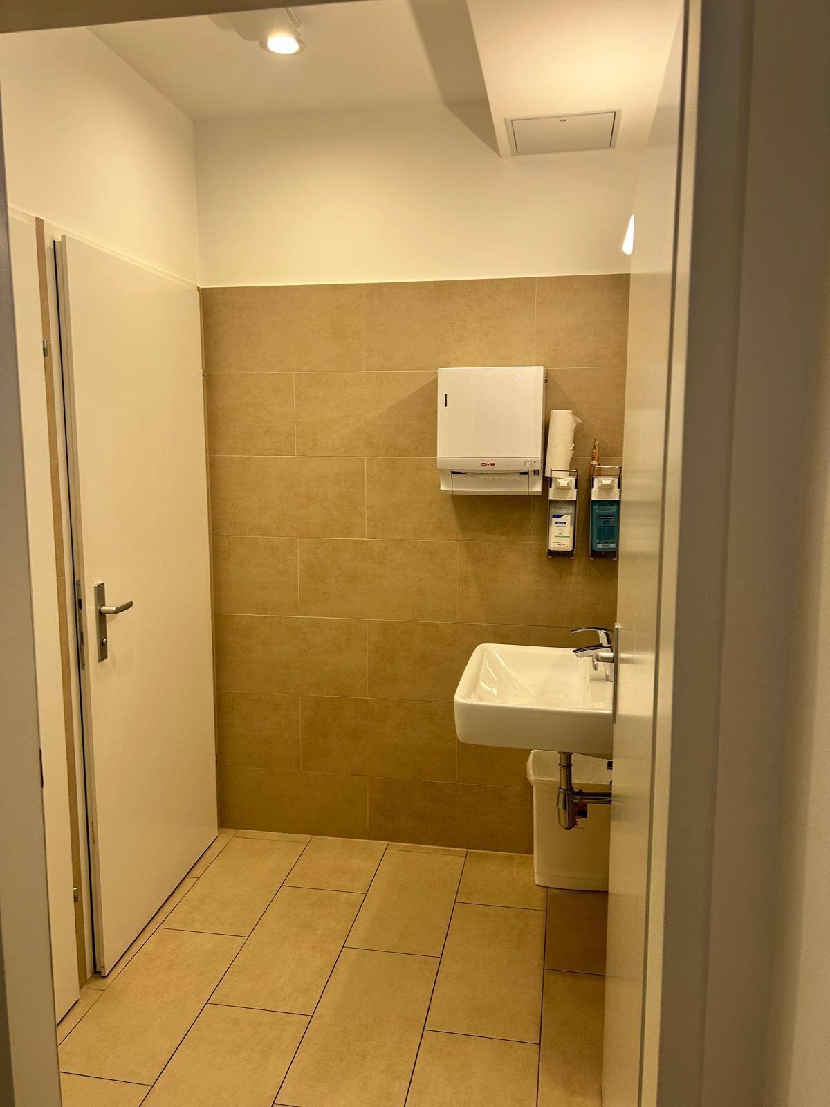 Saubere Toilette mit Waschbecken, beigen Fliesen und einem Spender für Papierhandtücher.