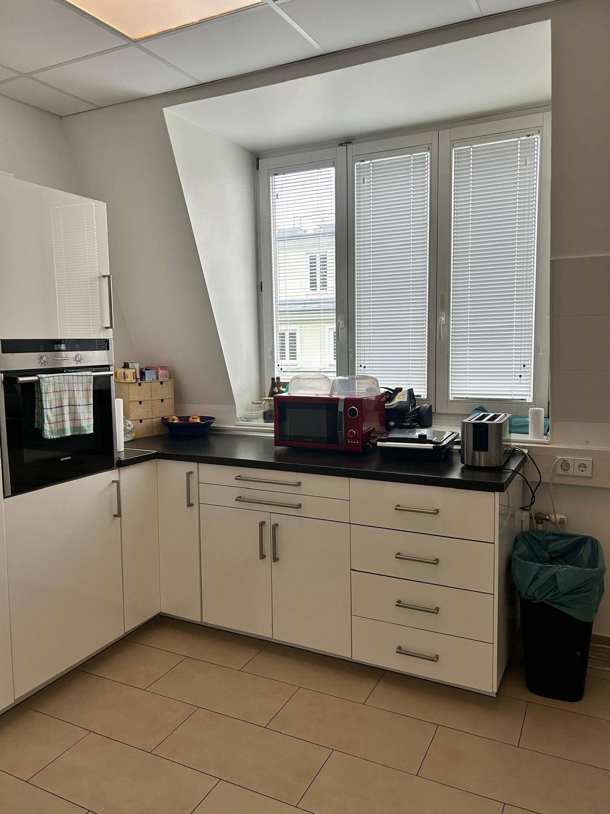 Kleine Büroküche mit weißen Schränken, dunkler Arbeitsplatte und Fliesenboden.