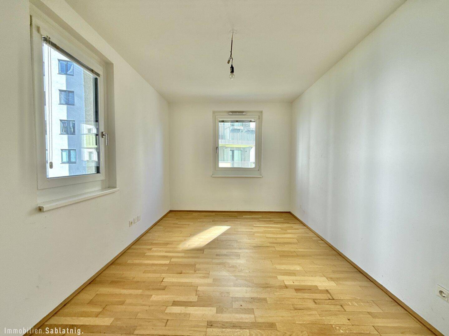 Helles, unmöbliertes Schlafzimmer mit Parkettboden und zwei Fenstern für viel Tageslicht.