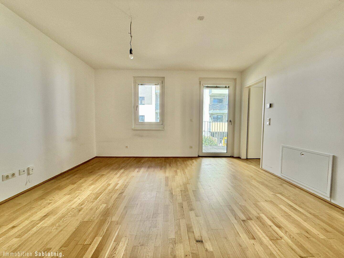 Geräumiges, unmöbliertes Wohnzimmer mit hellem Parkettboden und Balkonzugang.