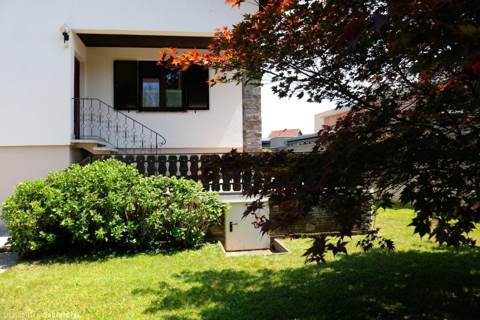 Eingangsbereich des Hauses mit kleiner Treppe und gepflegtem Vorgarten mit Bäumen und Sträuchern.