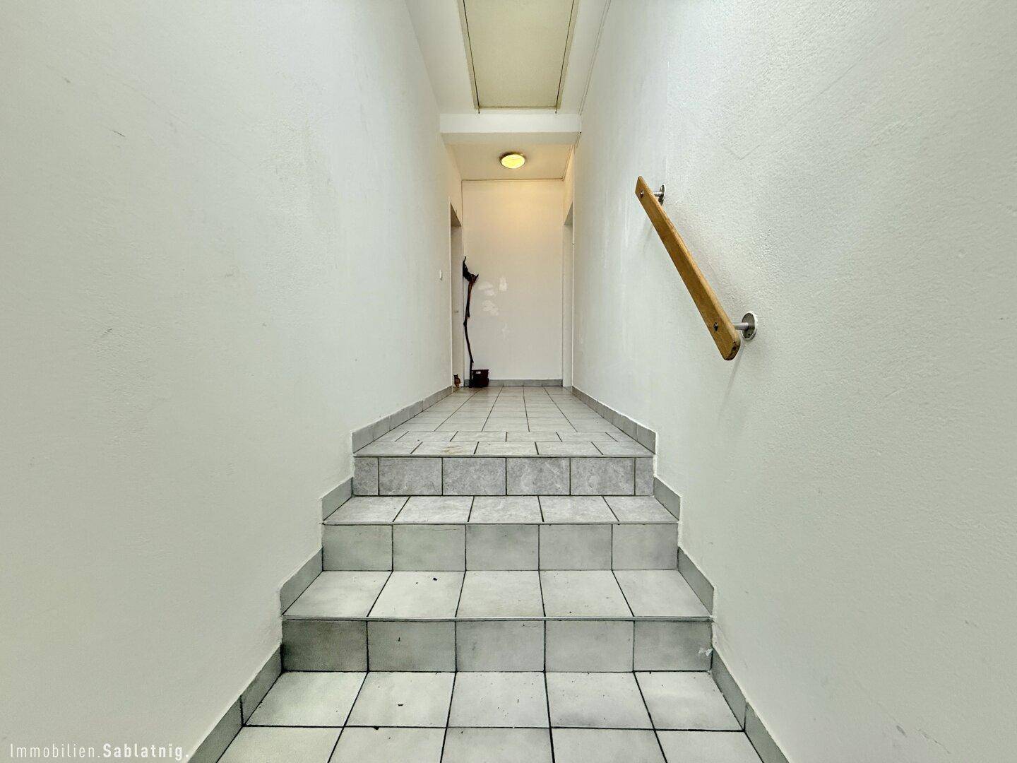 Heller Treppenaufgang mit Fliesenboden und einem hölzernen Handlauf an der Wand.