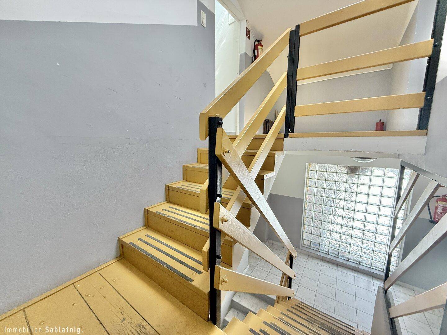 Treppenhaus mit gelben Holzstufen, Geländer und einer Glasbausteinwand für natürliches Licht.