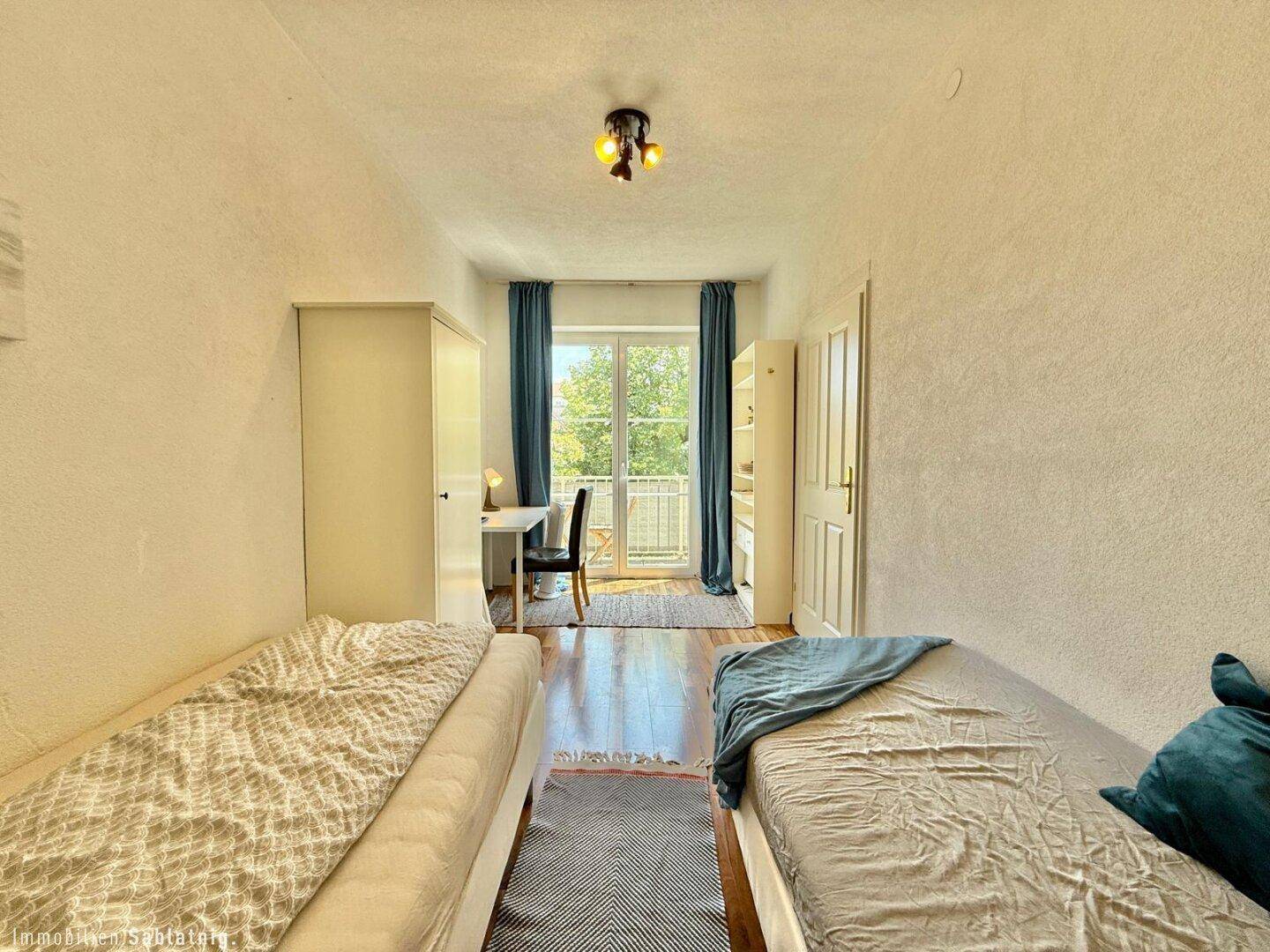 Schlafzimmer mit zwei Einzelbetten, Schreibtisch und Zugang zum Balkon.