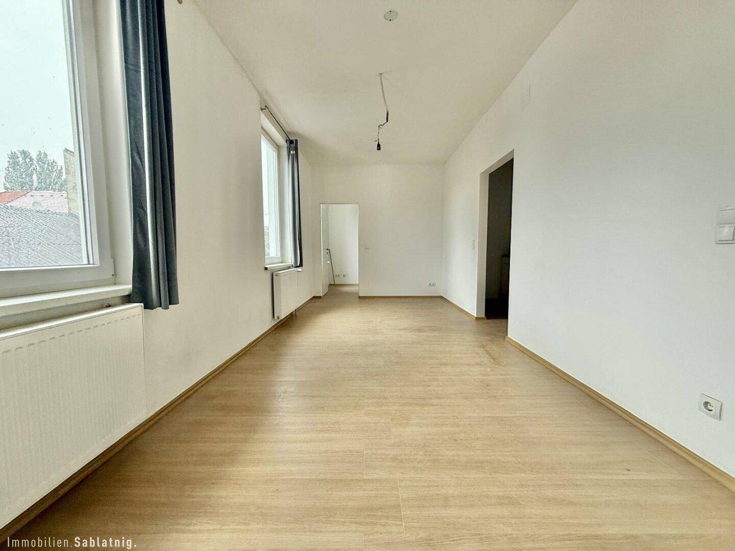 Geräumiges, helles Zimmer mit Holzfußboden und großen Fenstern, bietet vielseitige Einrichtungsmöglichkeiten.