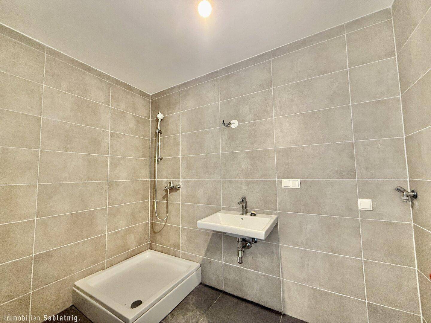 Modernes Badezimmer mit grauen Fliesen, ebenerdiger Dusche und wandhängendem Waschbecken.