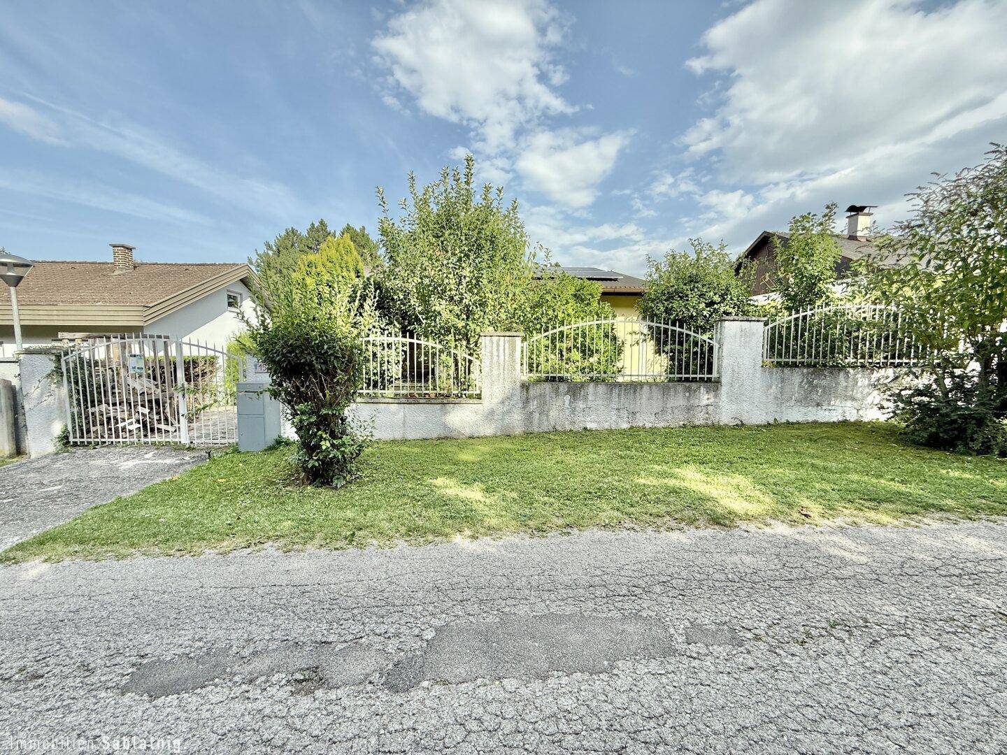 Vorgarten mit Rasenfläche, Zaun und dem Haus im Hintergrund unter blauem Himmel.