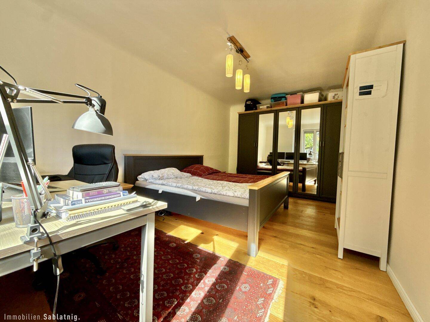 Schlafzimmer mit Holzboden, großem Bett und Arbeitsbereich am Fenster.