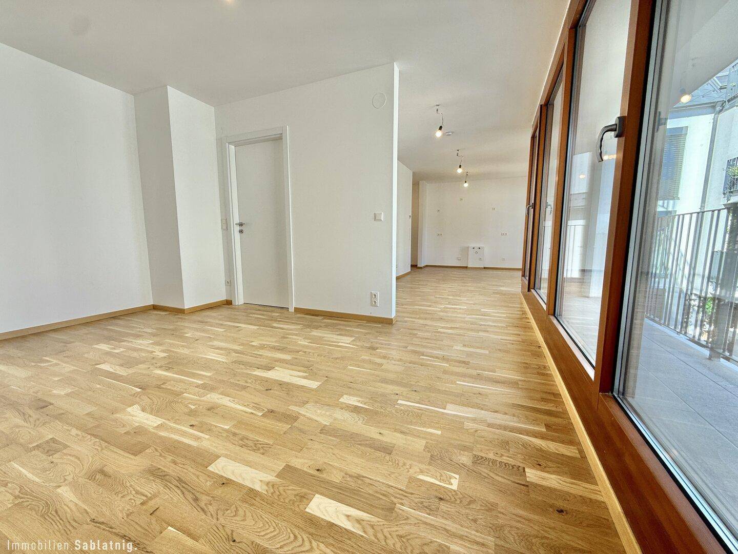 Geräumiger, leerer Raum mit Holzboden und breiter Fensterfront zum Außenbereich, lichtdurchflutet.