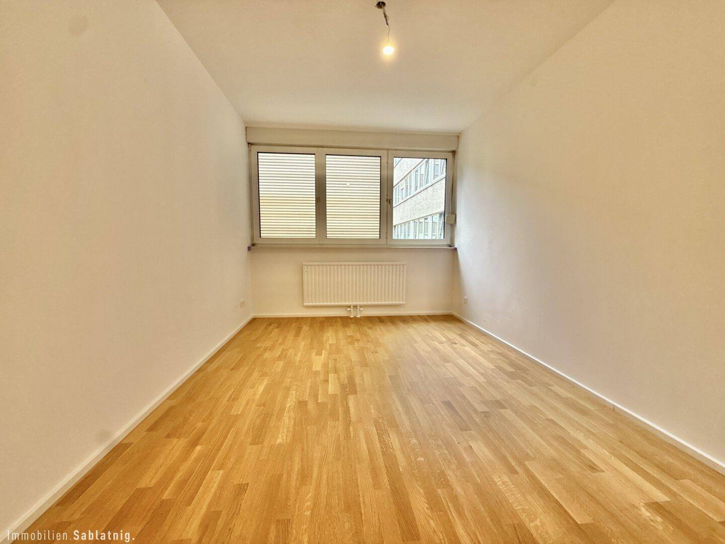 Leeres Schlafzimmer mit Holzboden und Fenster mit Blick auf die umliegenden Gebäude.