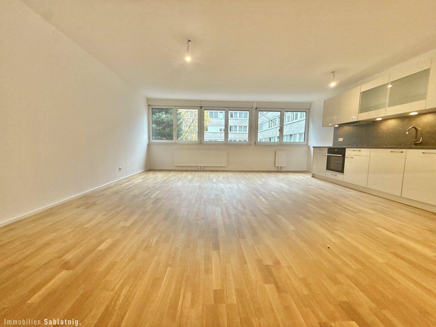 Großer, leerer Wohnbereich mit integrierter Küchenzeile, Holzboden und breiter Fensterfront.