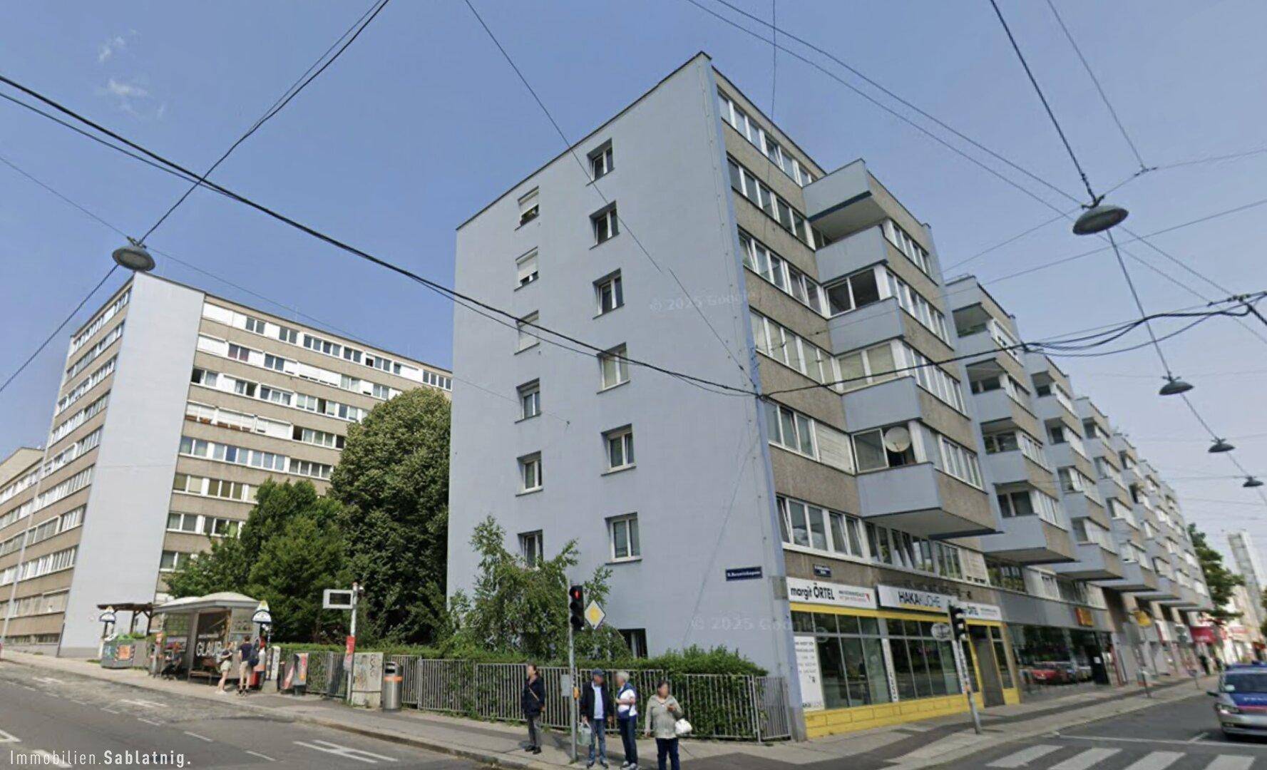 Mehrstöckiges Wohngebäude mit blauer Fassade und Geschäften im Erdgeschoss an einer Stadtstraße.