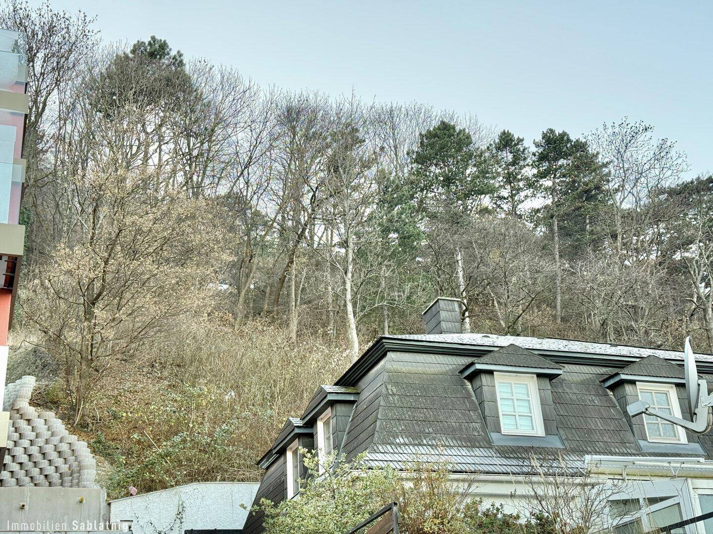 Gebäude mit Mansarddach und Gaubenfenstern, umgeben von Bäumen und einem bewachsenen Hang.