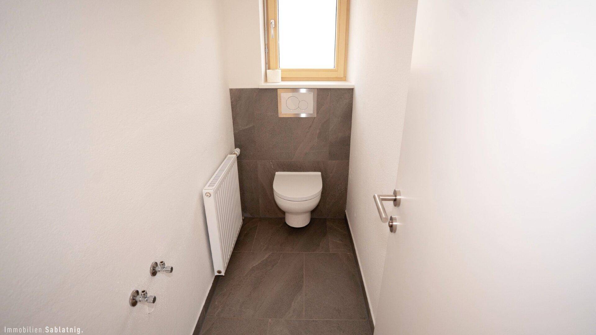 Modernes Gäste-WC mit Wand-WC, grauen Fliesen und Fenster für Tageslicht.
