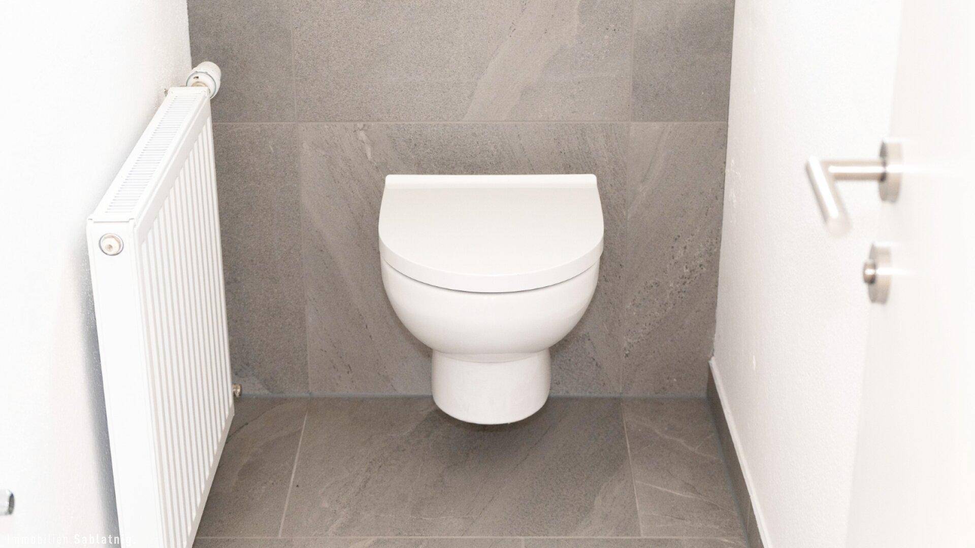 Wandmontiertes WC in einem modernen Badezimmer mit grauen Fliesen und Heizkörper.