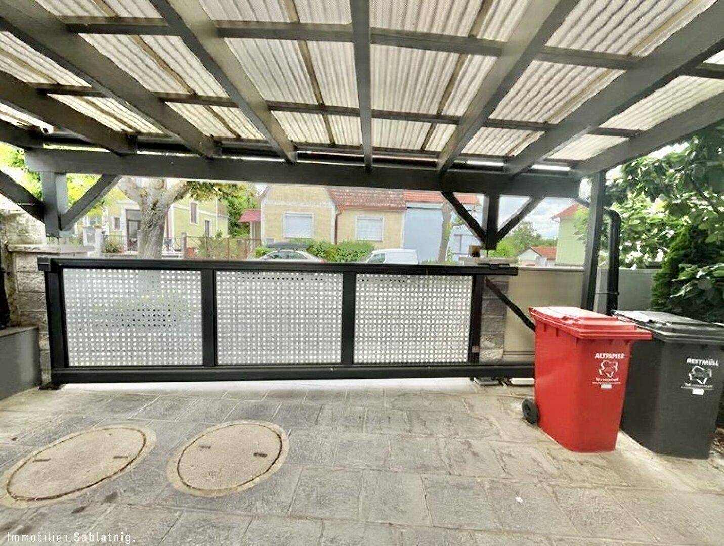 Überdachter Parkplatz mit Wellblechdach und Schiebetor zur Straße.