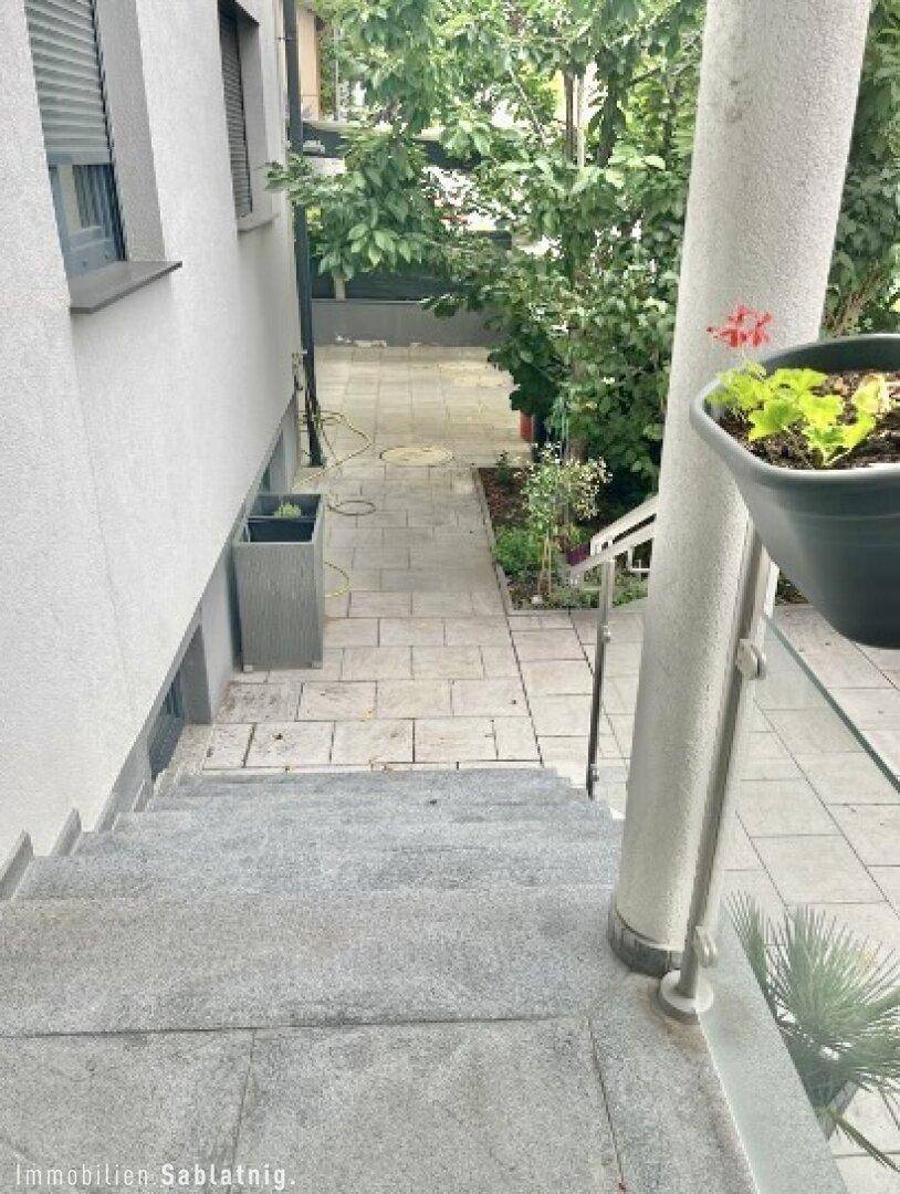 Seitlicher gepflasterter Weg entlang des Hauses mit Pflanzen und Treppenzugang.
