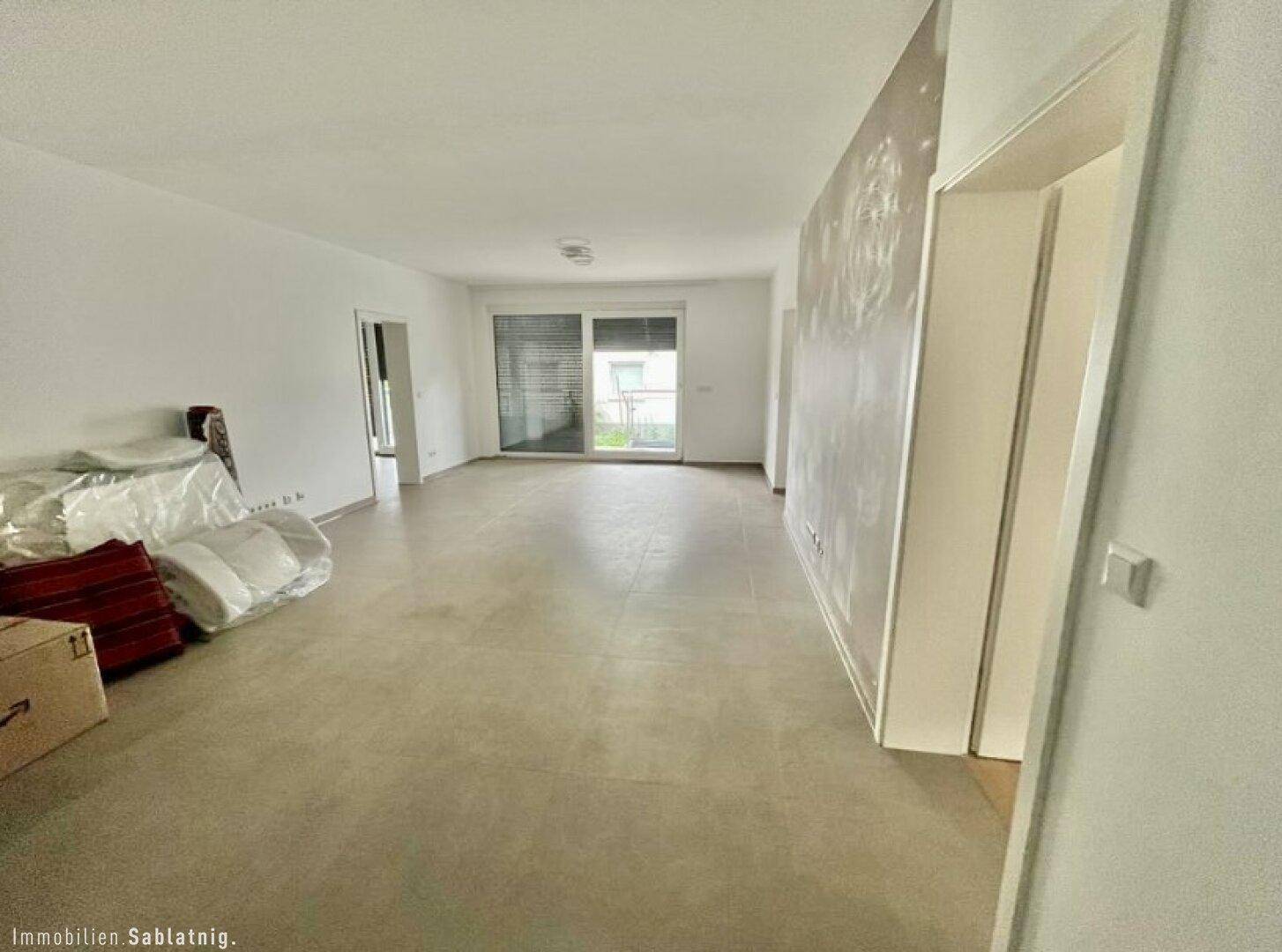 Leeres Wohnzimmer mit Fliesenboden und Zugang zur Terrasse durch große Schiebetüren.
