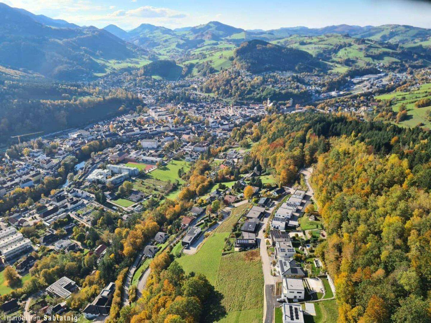 Luftaufnahme einer Stadt in einem Tal, umgeben von Bergen und herbstlich gefärbten Wäldern.