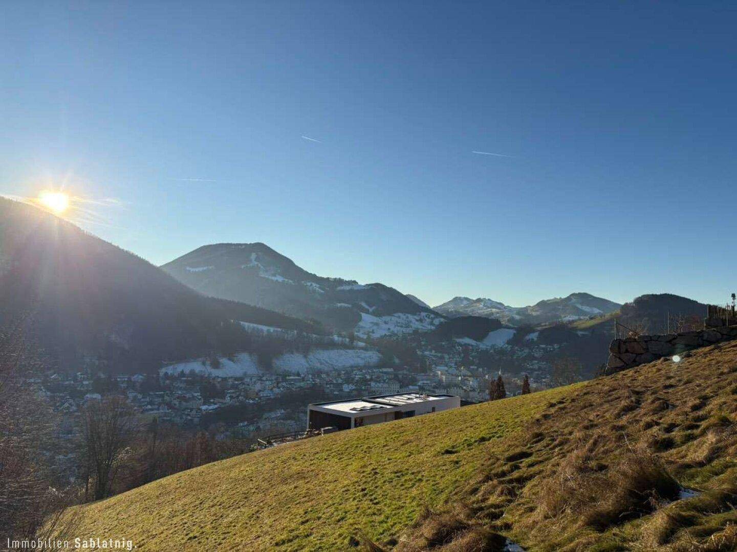 Sonnige Berglandschaft mit modernem Gebäude auf einem grünen Hang und weitem Blick auf die Stadt.