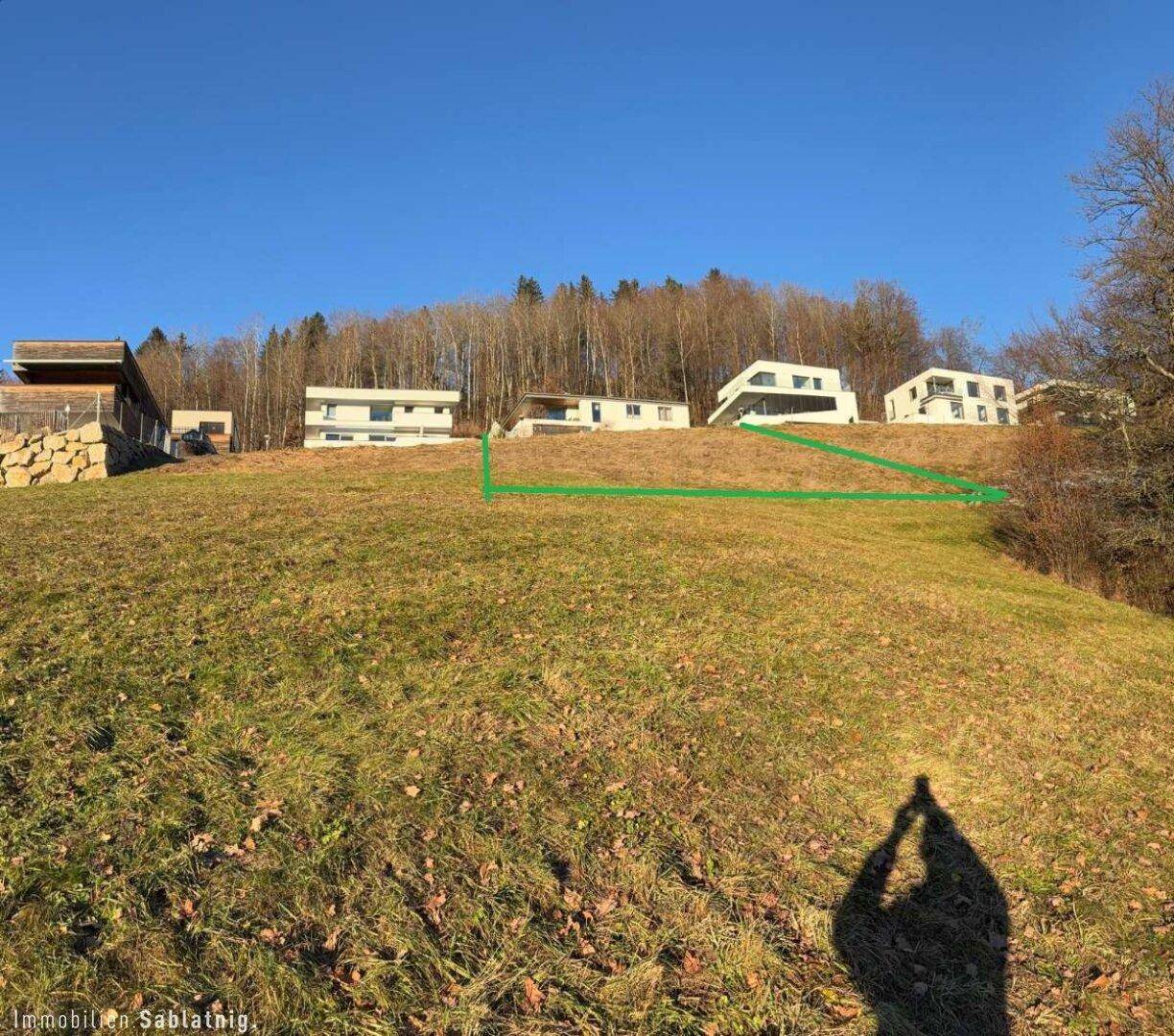 Grüner Hang mit modernen Gebäuden im Hintergrund, umgeben von Wald und mit sonnigem Ausblick.